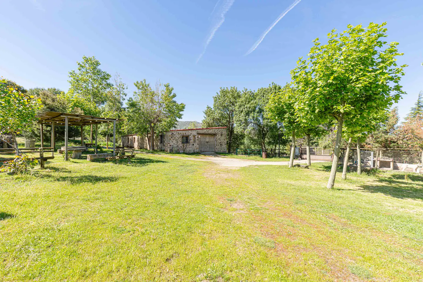 Outdoor shot of a stone building with a wooden door, surrounded by green grass, trees, and a picnic area under a blue sky.