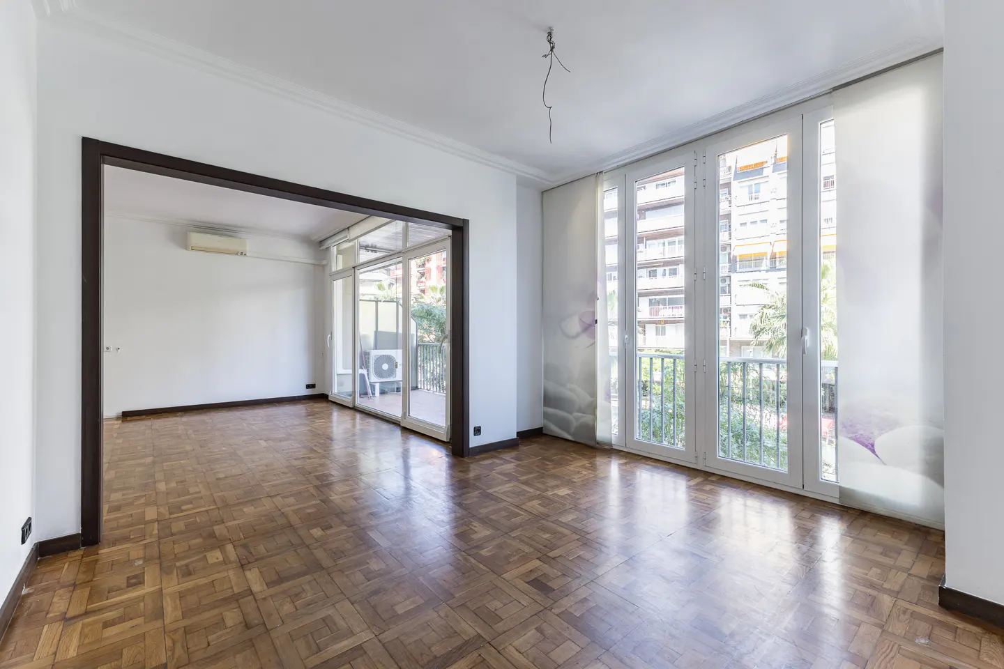 Bright, empty room with parquet floors, white walls, and large windows overlooking a balcony and city view. Dark wood trim frames an adjacent room.