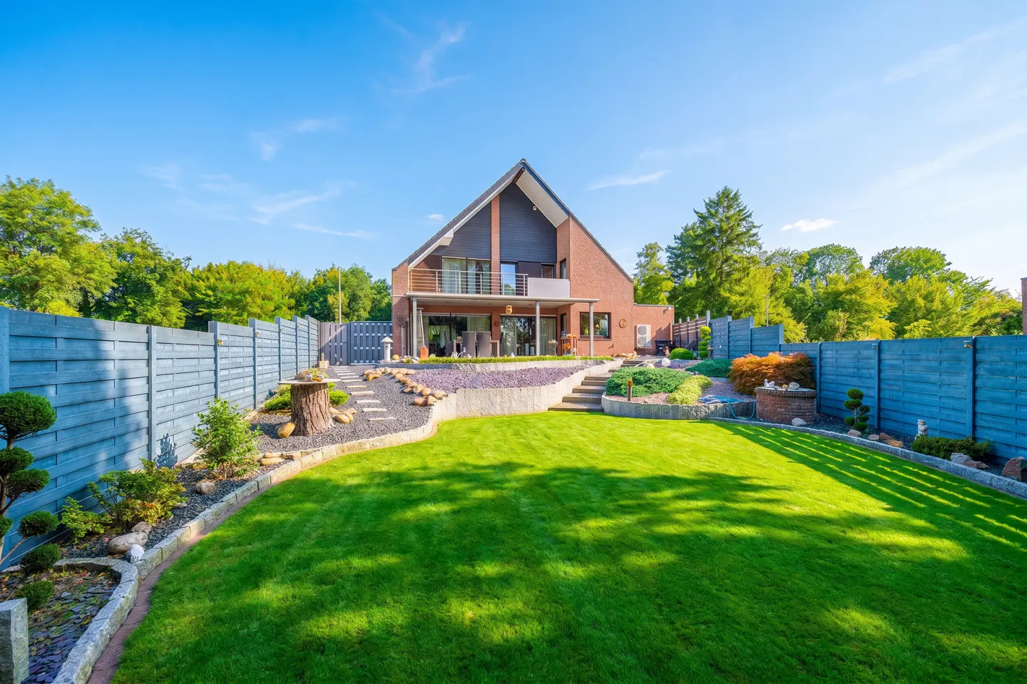 A modern brick house with a green lawn and blue fence under a blue sky. The house has a balcony and a patio.