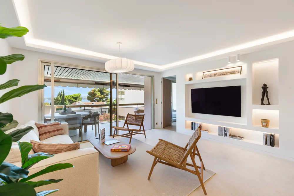 Modern living room with white walls, a beige sofa, two rattan chairs, a wall-mounted TV, and a balcony with sea views.