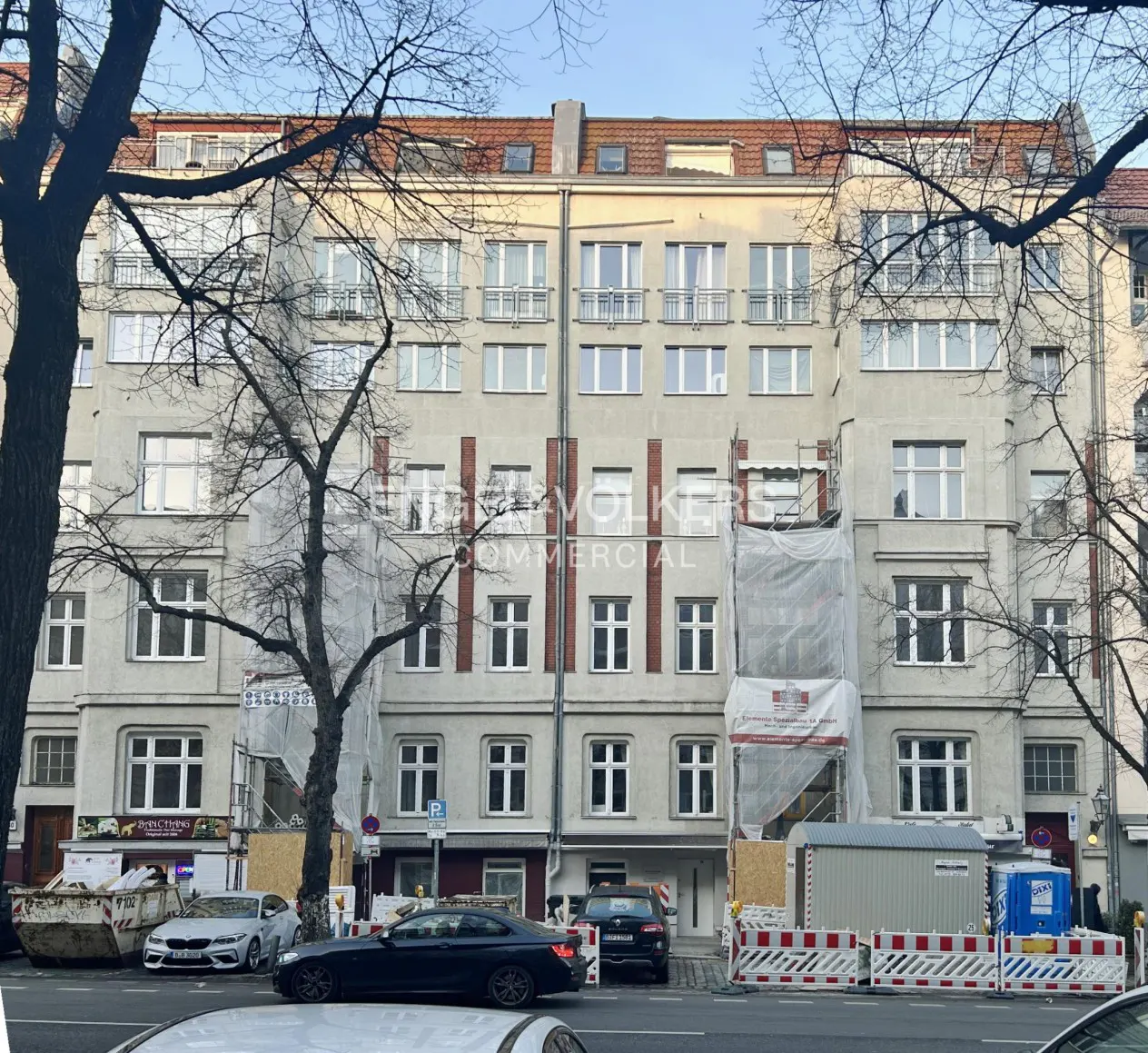A five-story apartment building with white windows and a red-tiled roof, partially obscured by bare trees and construction barriers.