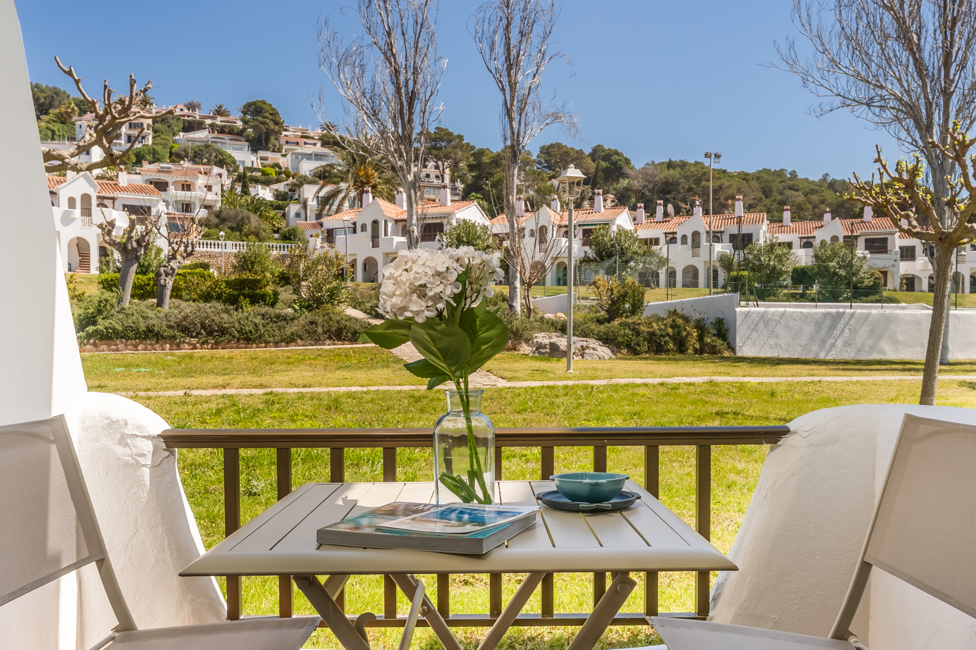 Balcony view of white houses with red roofs. Table with flowers, book, and bowl. Green grass and trees in the background.