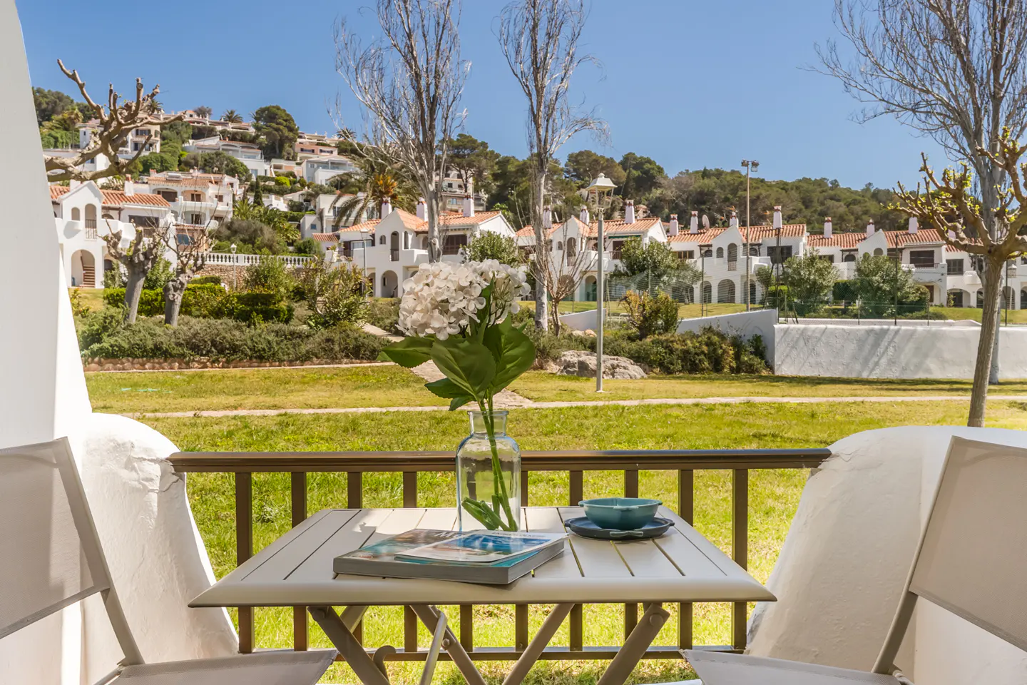 Balcony view of white houses with red roofs. Table with flowers, book, and bowl. Green grass and trees in the background.