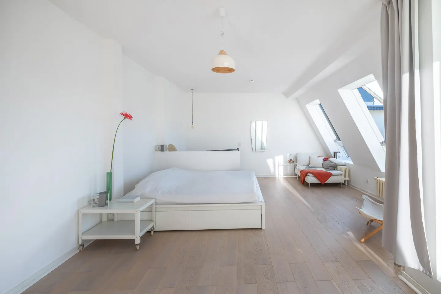 Bright, modern bedroom with white walls, light wood floors, and skylights. A bed, sofa, and side table furnish the space. A red flower adds a pop of color.