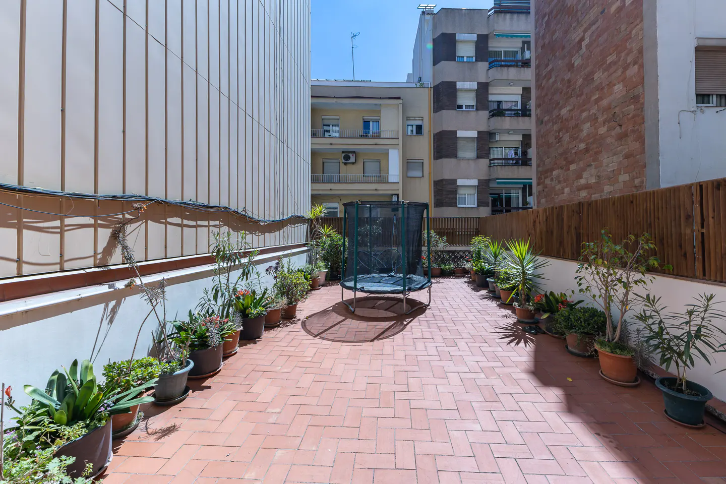 Outdoor patio with brick flooring, potted plants, and a trampoline. Buildings are visible in the background.