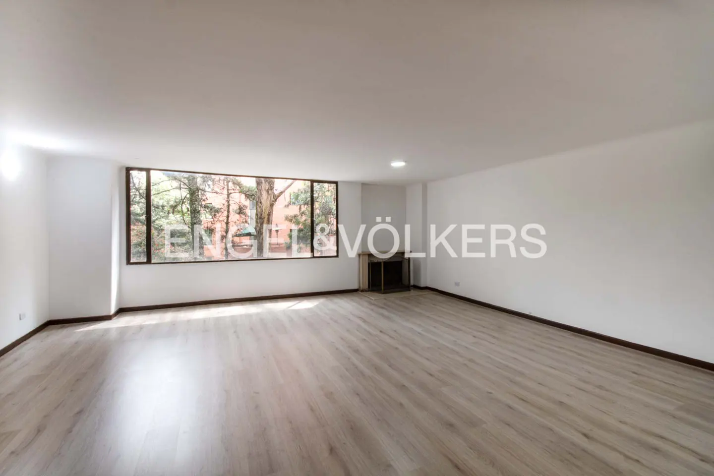 Spacious, empty living room with light wood floors, white walls, and a large window overlooking trees.