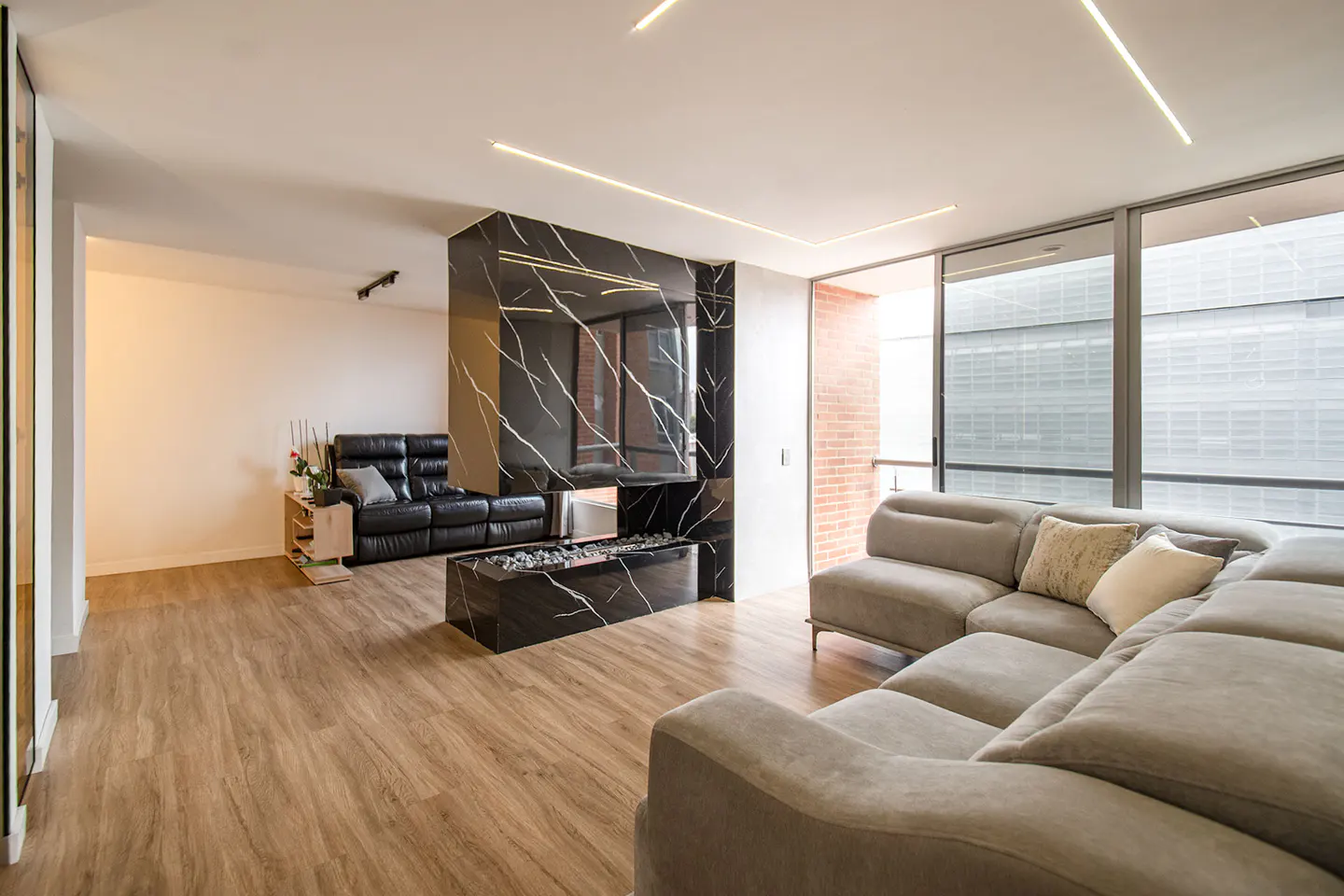 Living room with wood floors, a gray sectional sofa, and a black marble fireplace. A black leather sofa is visible in the background.