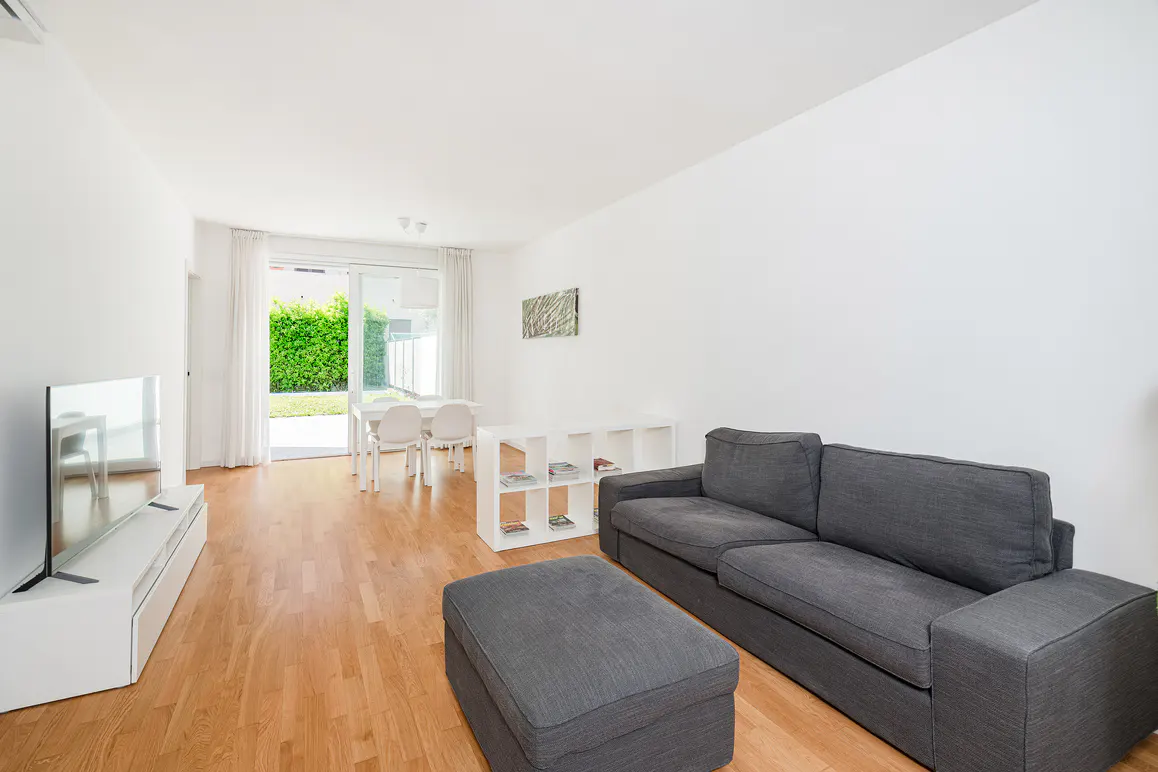 Bright living room with hardwood floors, white walls, and a gray sofa. A white table and chairs are visible through the open patio door.