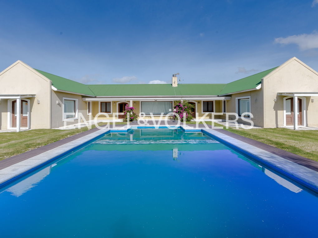 A U-shaped beige house with a green roof surrounds a blue swimming pool under a blue sky.