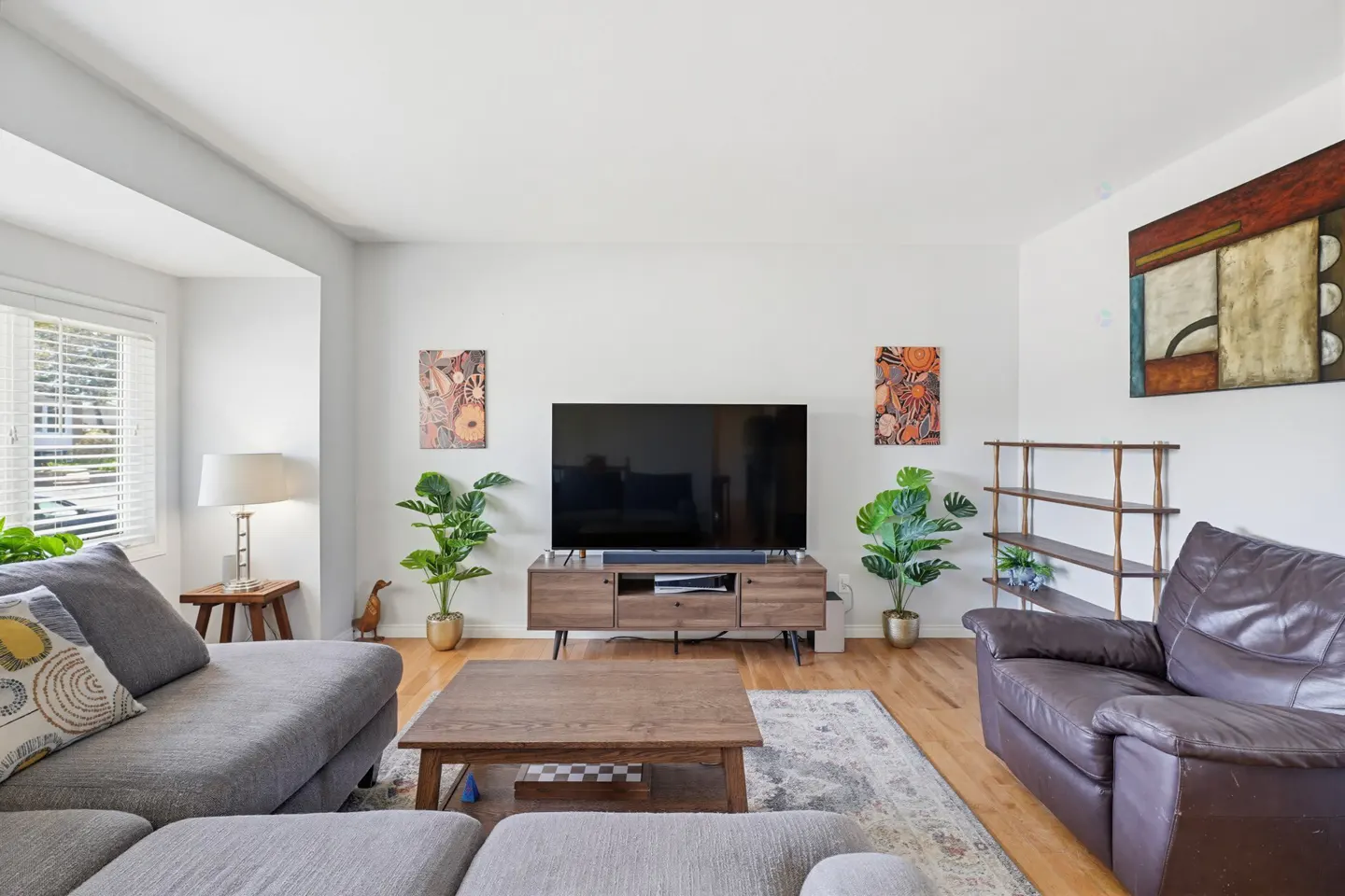 Modern living room with a grey sectional, brown leather armchair, wooden coffee table, TV console, and decorative art.