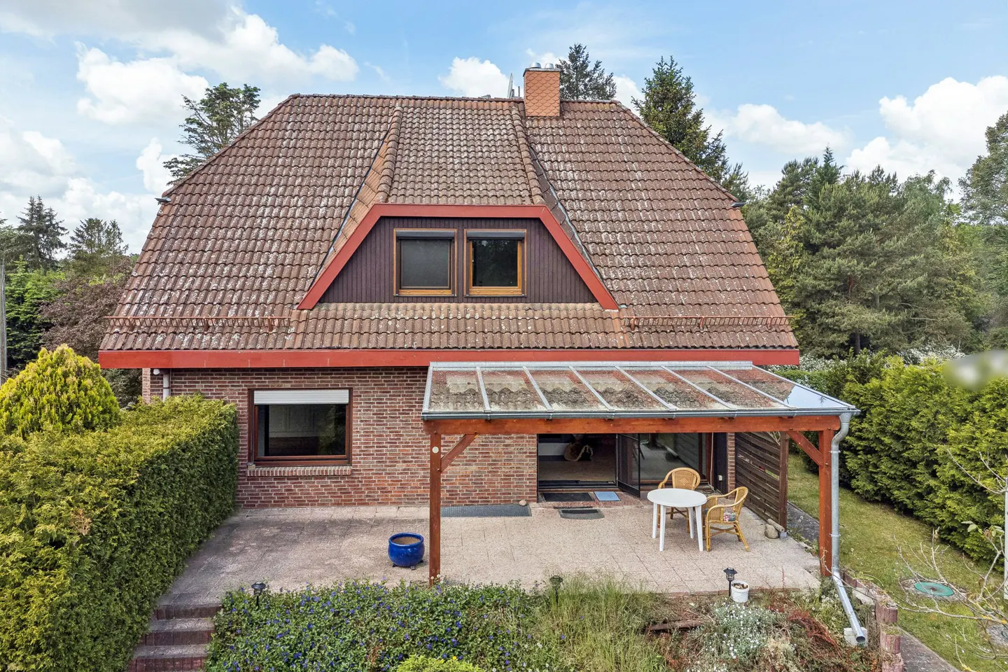 Two-story brick house with a red tile roof and a covered patio with a table and chairs. Green trees surround the house.