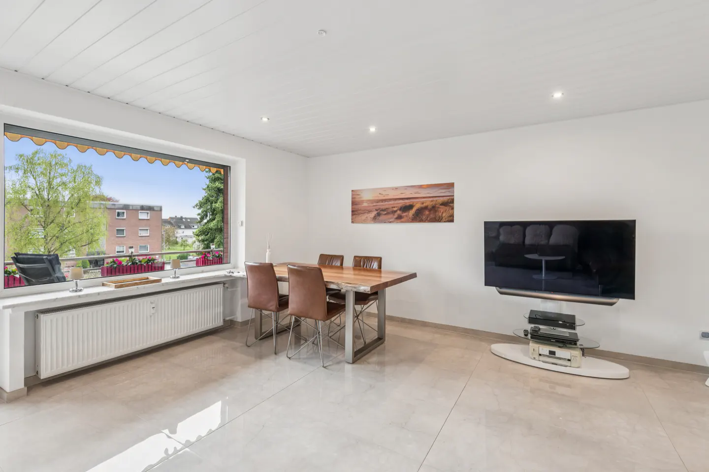 Bright living room with a wood dining table, leather chairs, and a large window with a view of trees and buildings. A flat-screen TV is mounted on the wall.