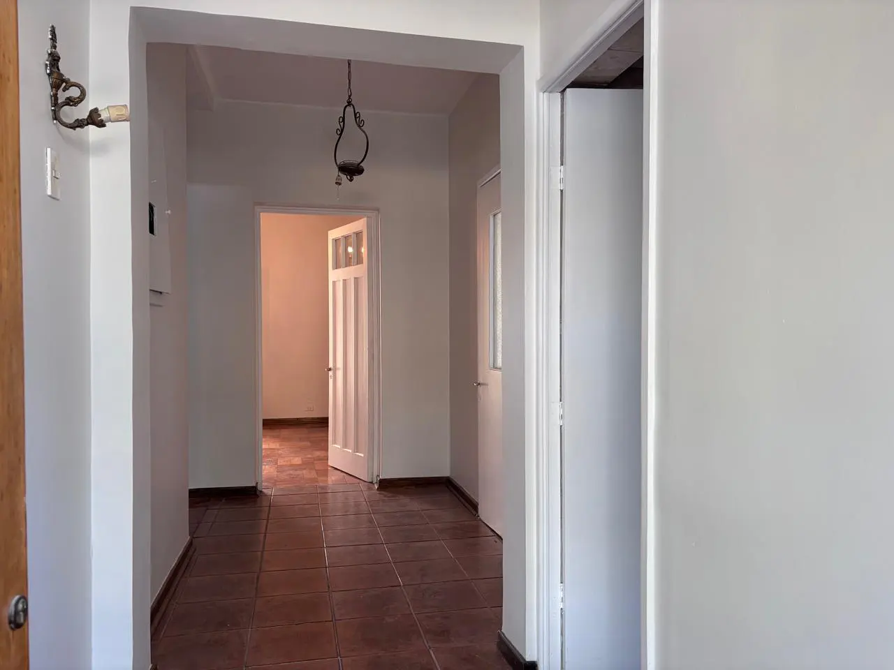 Hallway with brown tile floor, white walls, and doorways leading to other rooms. A vintage light fixture hangs from the ceiling.