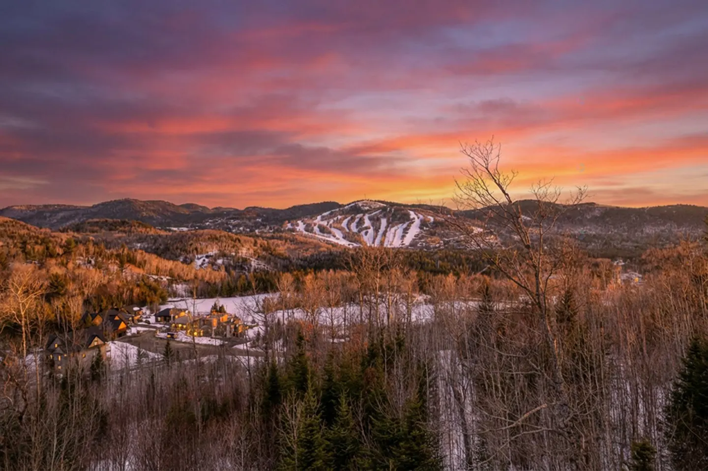 Winter landscape view of mountains and ski resort at sunset with orange and pink sky. Houses nestled in snowy valley.