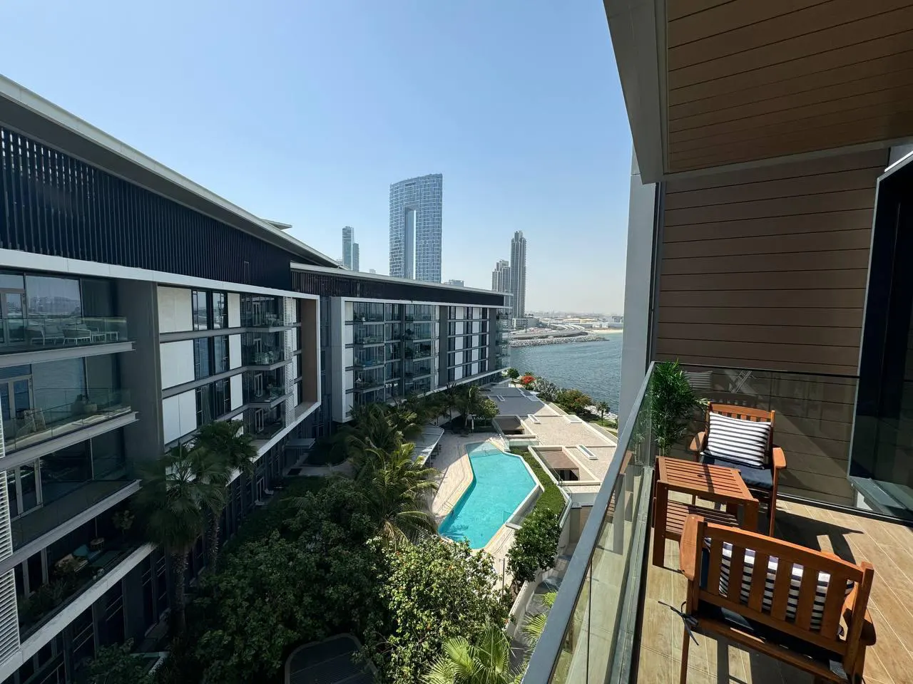 Balcony view of a modern apartment complex with a pool, palm trees, and city skyline in the background. Wooden furniture on the balcony.