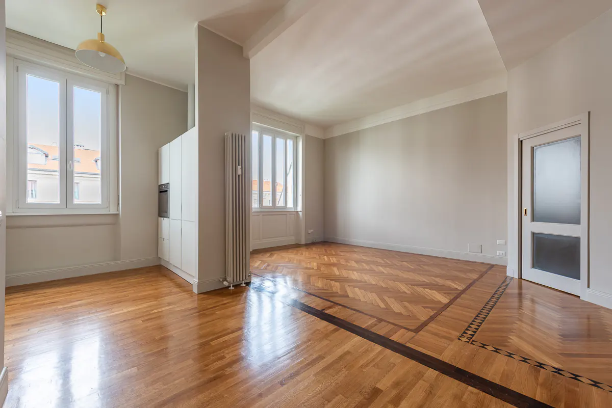 Bright, empty room with herringbone wood floors, white walls, and large windows. A white cabinet and radiator are visible.