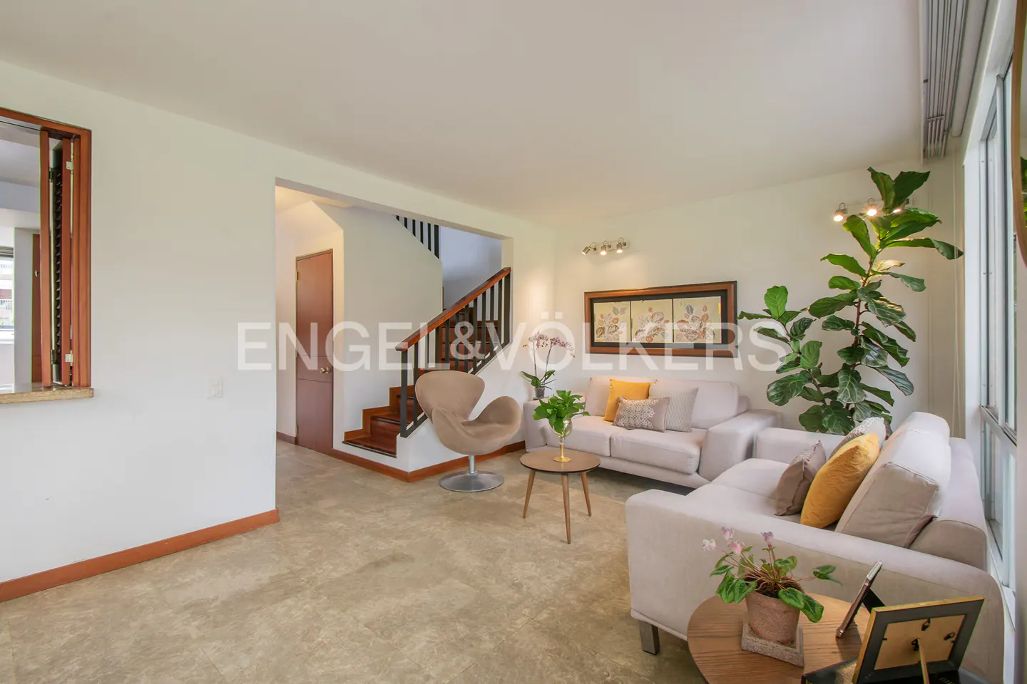 Living room with white walls, beige sofas, a tan chair, and a staircase in the background. A large plant sits by the window.