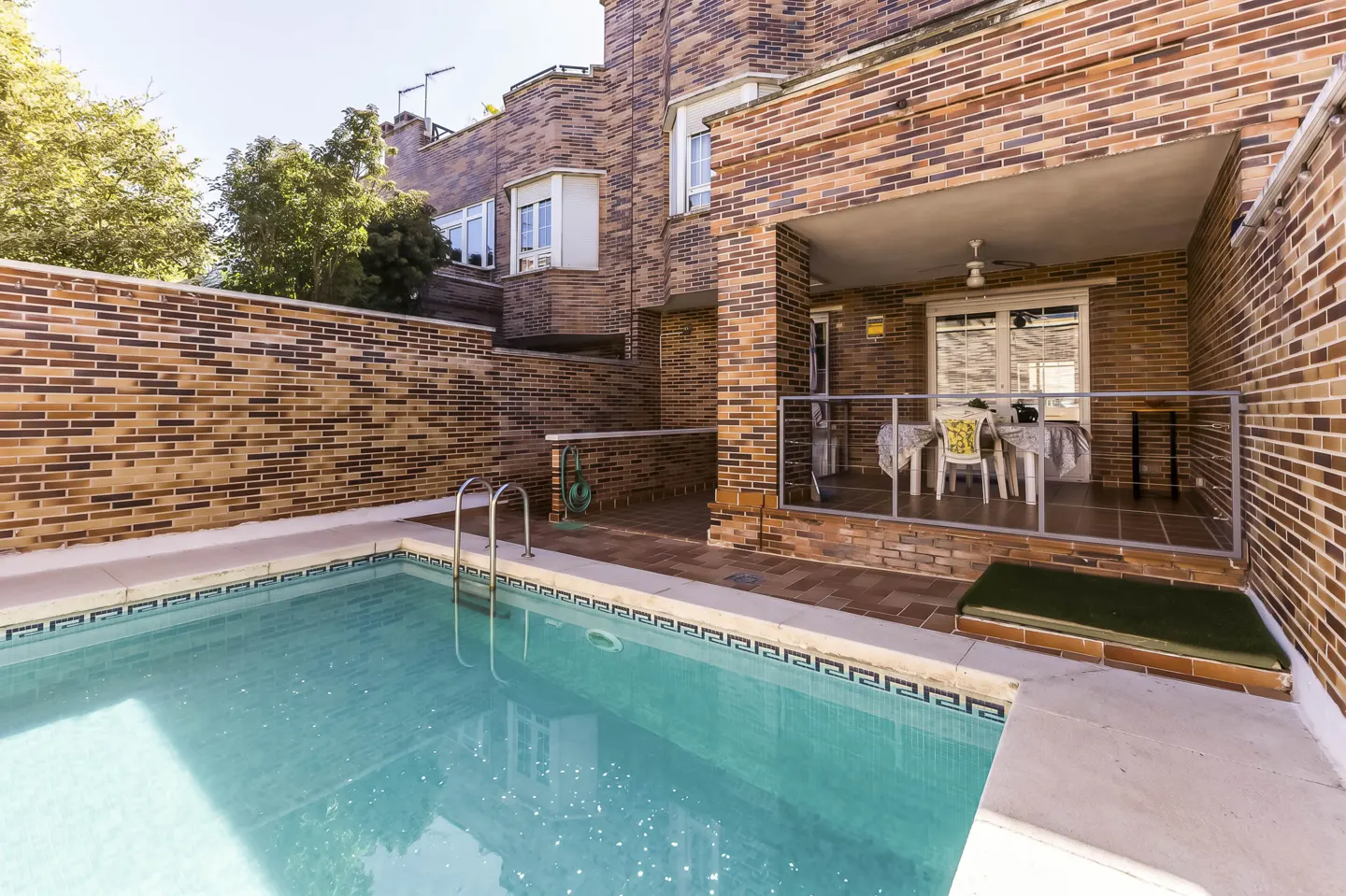 A backyard with a pool and a brick house. The pool has a ladder and the house has a covered patio with a table and chairs.