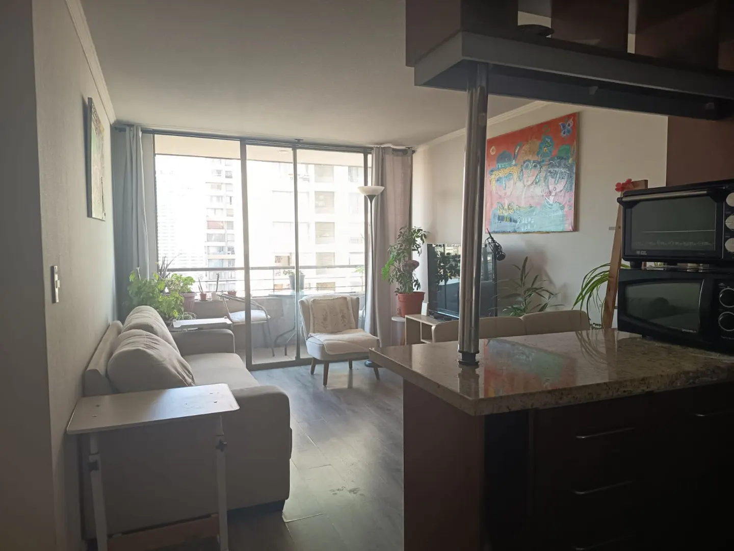 Living room with a gray sofa, chair, and balcony with city views. A kitchen island with granite countertop is in the foreground.