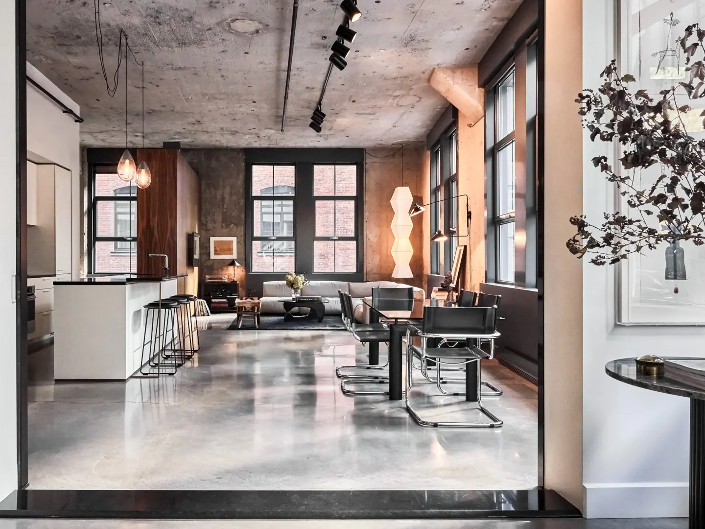 Open-concept loft with concrete floors, exposed ceiling, and large windows. A white kitchen island and dining area are visible.