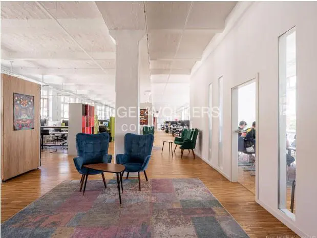 Open office space with wood floors, white walls, and exposed ceiling. Two blue chairs sit on a colorful rug. Desks and people are visible in the background.