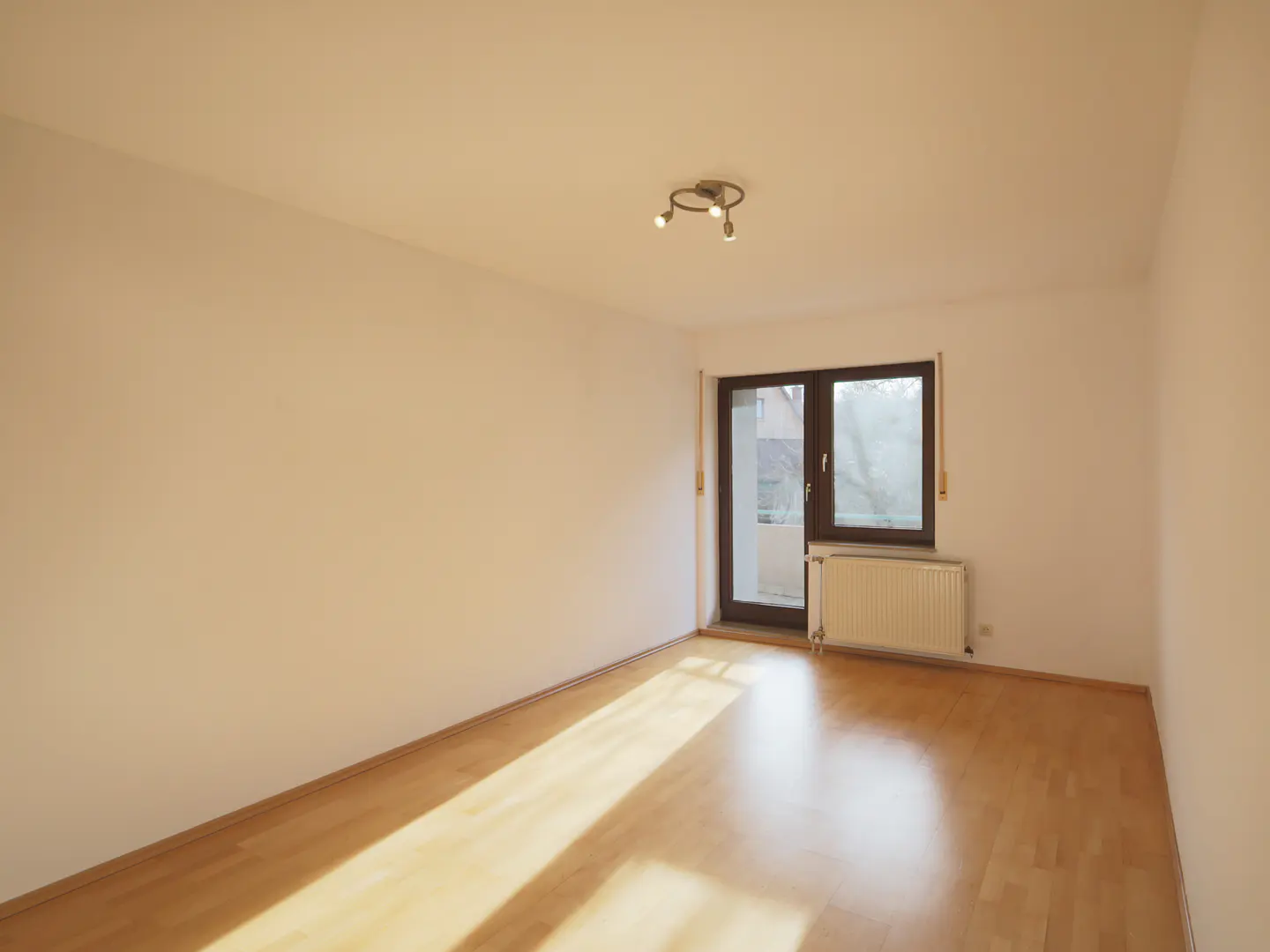 Empty room with light wood floors, cream walls, and a window with a radiator below. A modern light fixture is on the ceiling.