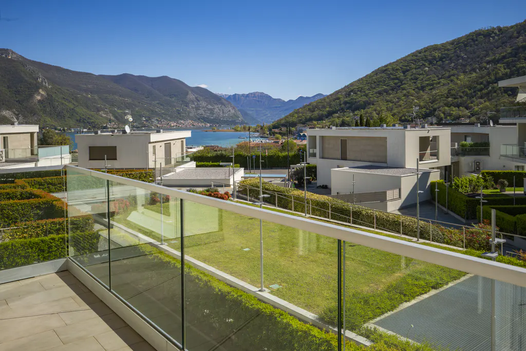 Balcony view of modern white houses, green lawns, and a lake surrounded by mountains under a clear blue sky.