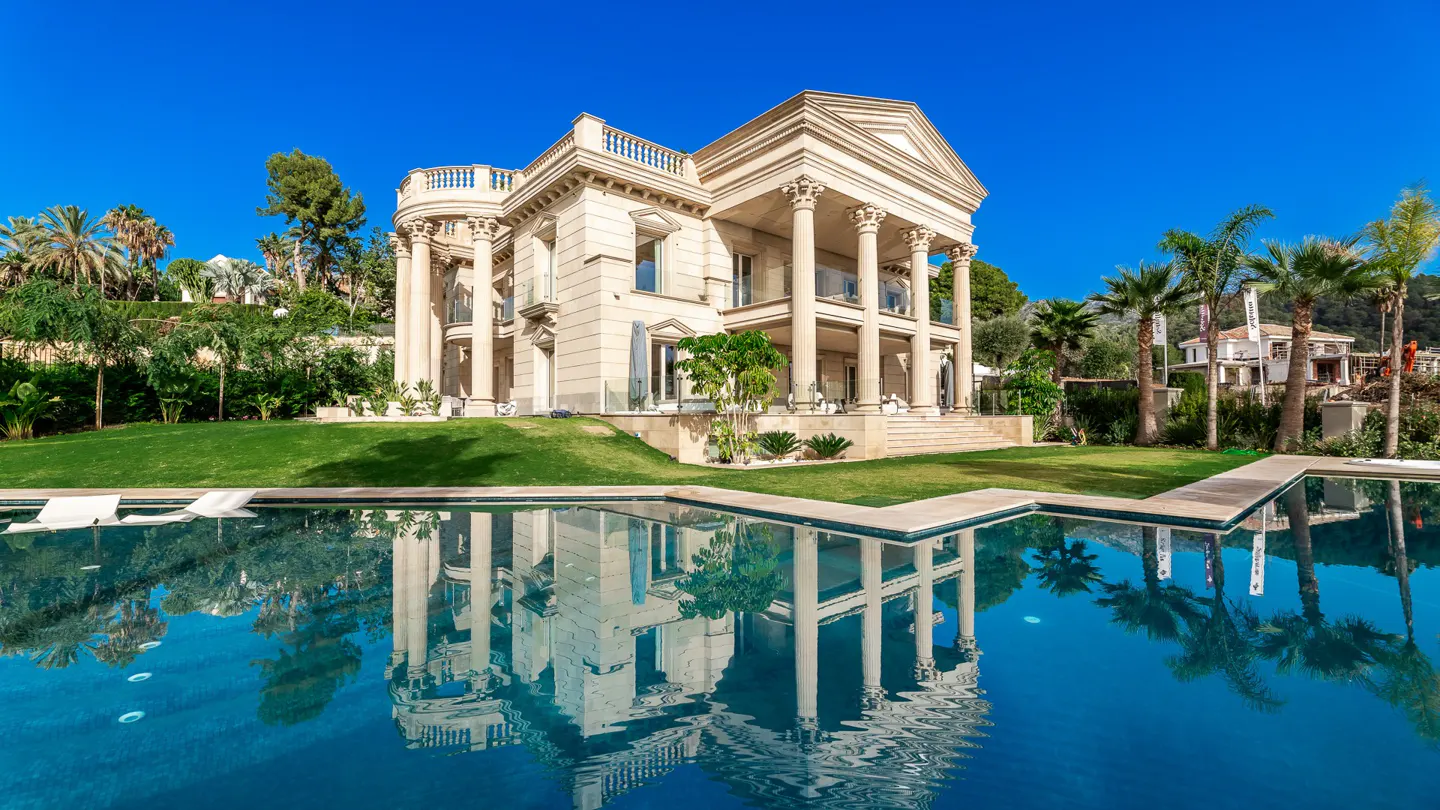 Beige mansion with columns reflected in a blue pool. Green lawn, palm trees, and clear blue sky.