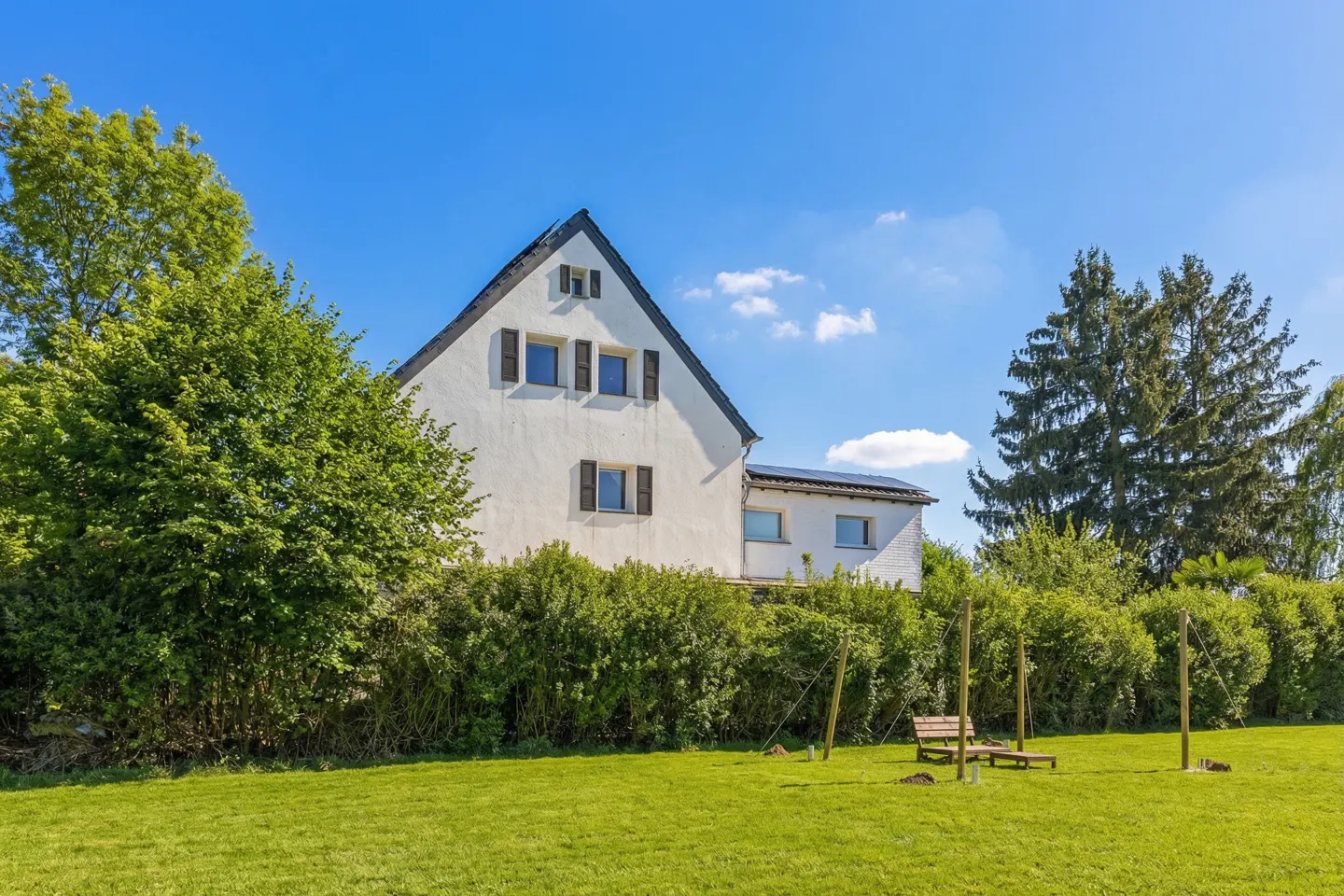 Two-story white house with dark shutters, green lawn, trees, and a swing set under a blue sky.