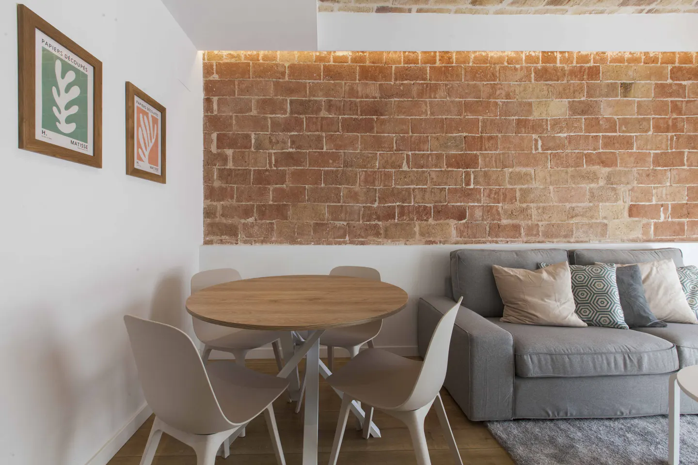A dining area with a round wooden table, four white chairs, and a grey sofa against an exposed red brick wall.