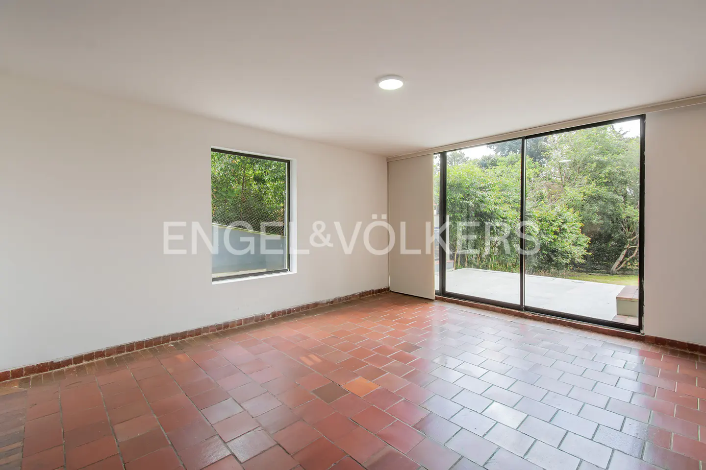 Empty room with red tile floor, white walls, and large windows overlooking green trees. Engel & Völkers logo visible.