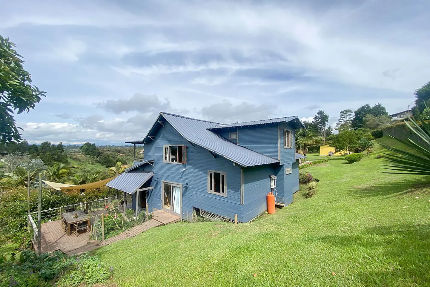 Two-story blue house with a metal roof on a green hillside under a cloudy sky. A wooden deck with a table is visible.