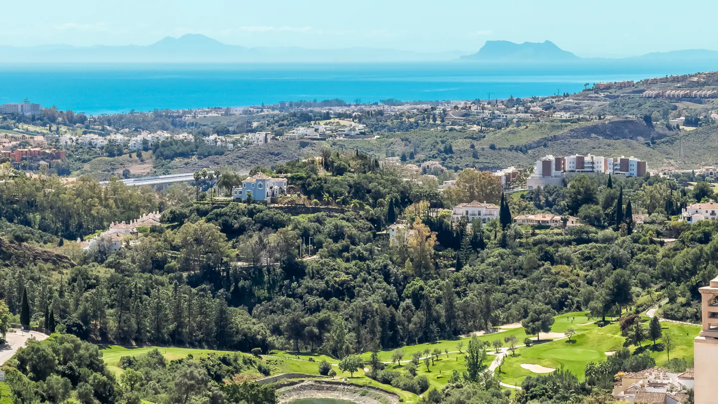 Scenic view of a lush, green hillside with scattered homes, a golf course, and the blue ocean in the background.