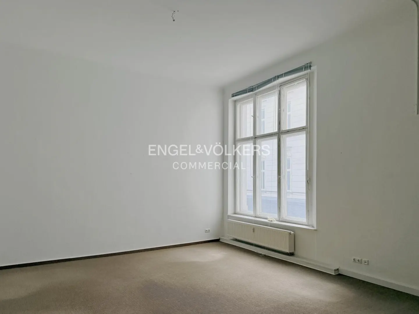 Empty room with white walls, brown carpet, and a large window with white frame. A radiator is under the window. Engel & Völkers logo on the wall.