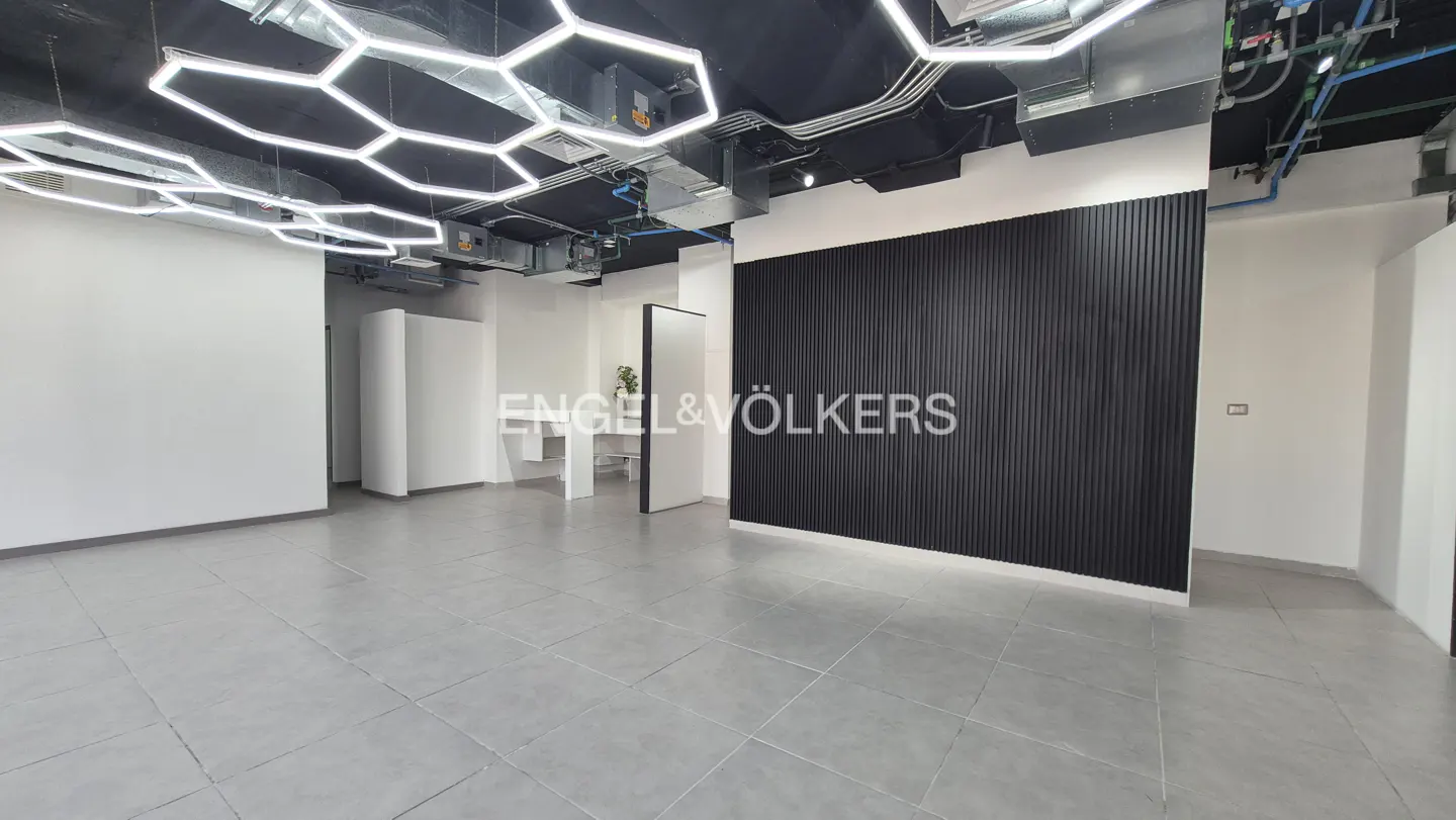 An empty office space with gray tile floors, white walls, and a black accent wall. Hexagonal lights hang from the black ceiling.