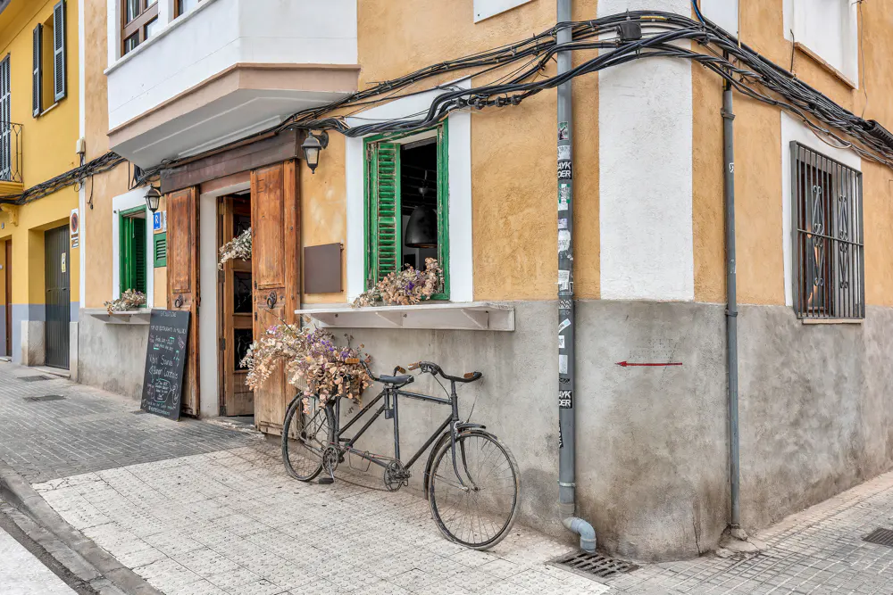 A bicycle with flowers in the basket is parked outside a yellow building with green shutters.