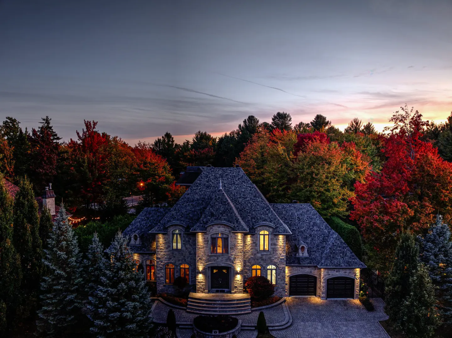 Stone house with lit windows at dusk, surrounded by colorful autumn trees.