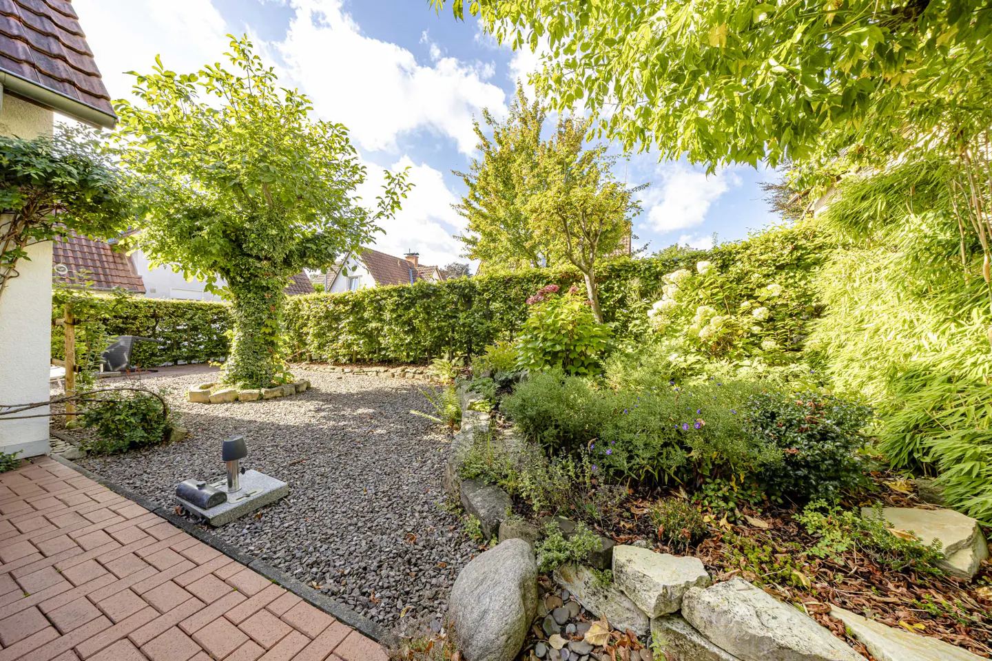 A lush garden with gravel path, brick patio, green hedges, trees, and various plants under a partly cloudy sky.
