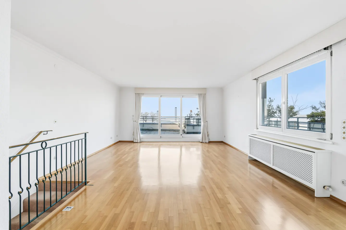 Bright, empty room with wood floors, white walls, large windows, and a balcony view. A black metal railing leads to stairs.
