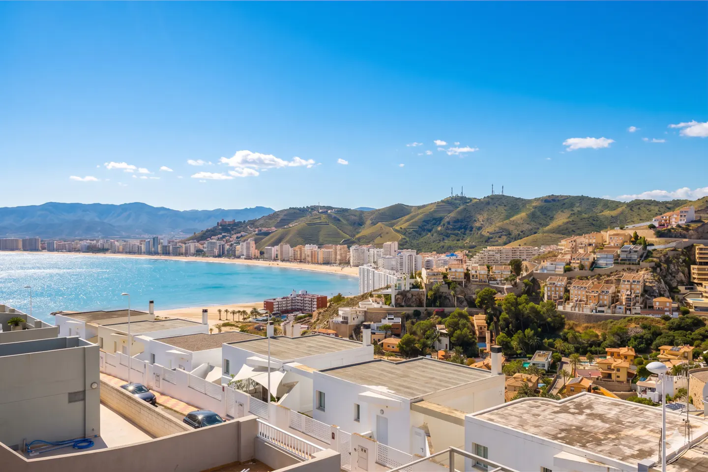 Scenic view of a coastal city with white buildings, a sandy beach, turquoise water, and green hills under a bright blue sky.