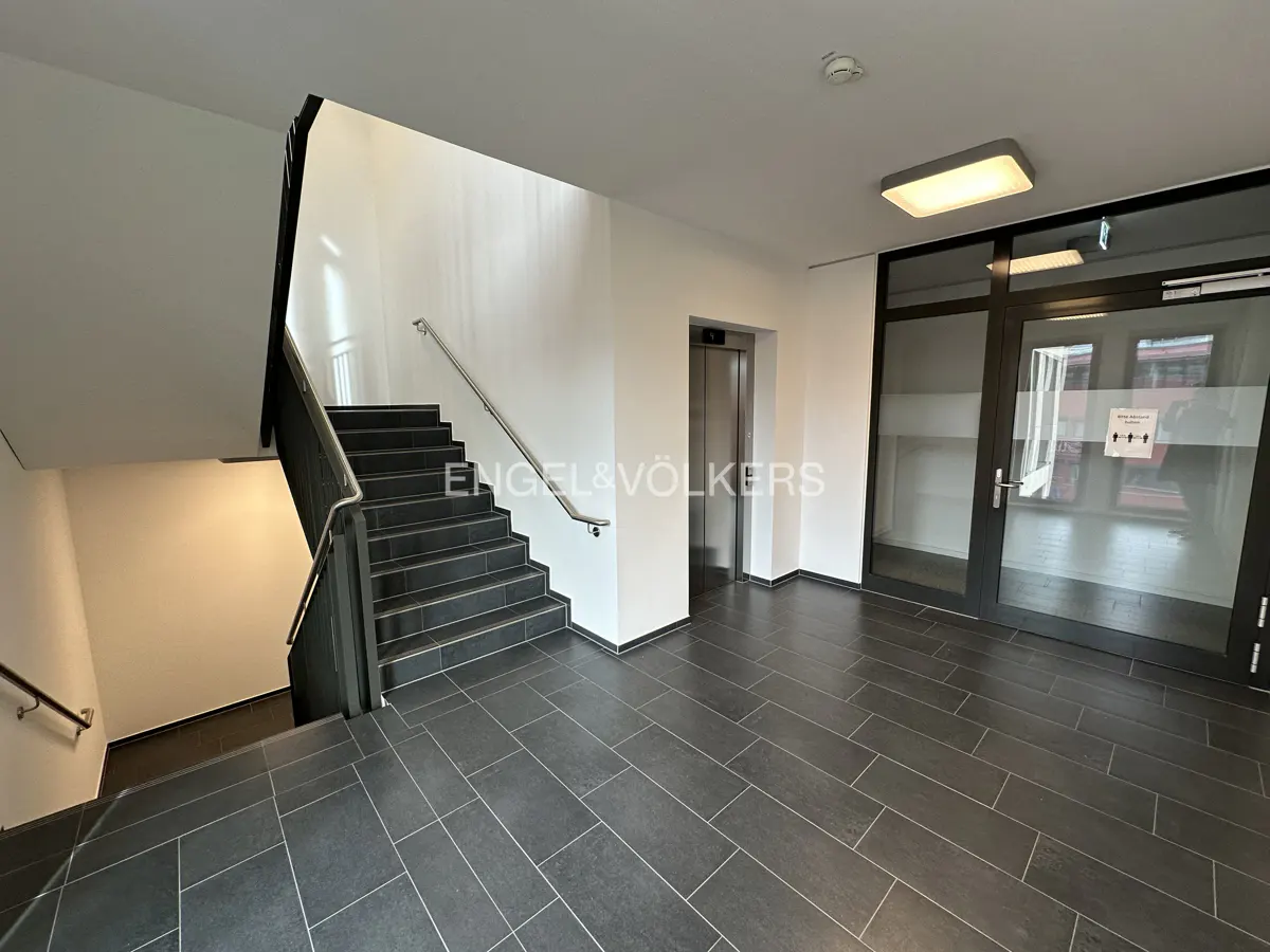A modern building lobby with black tile floors, white walls, a staircase, an elevator, and glass doors.
