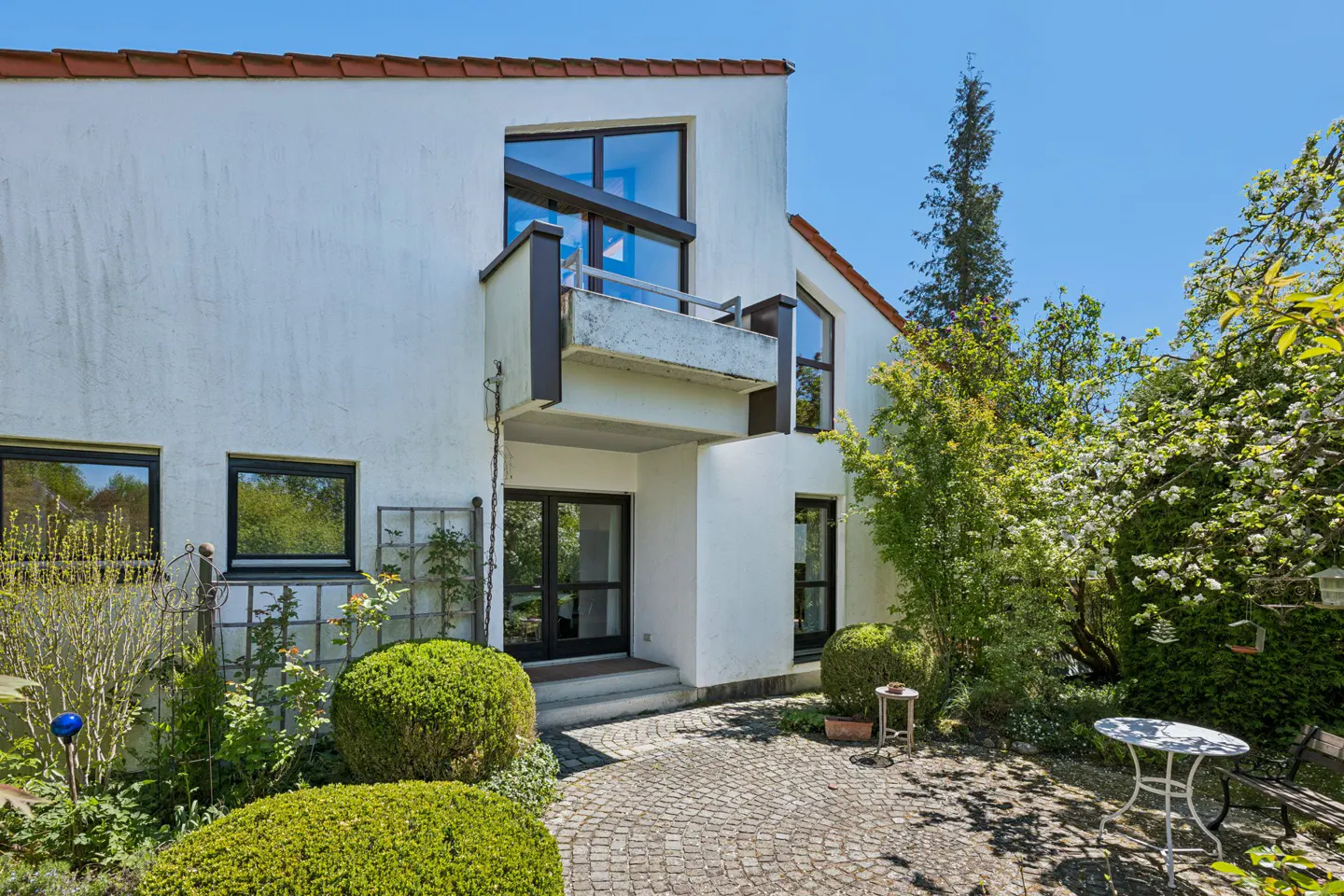 Exterior view of a white two-story house with a balcony, black framed windows, and a cobblestone patio surrounded by green bushes.