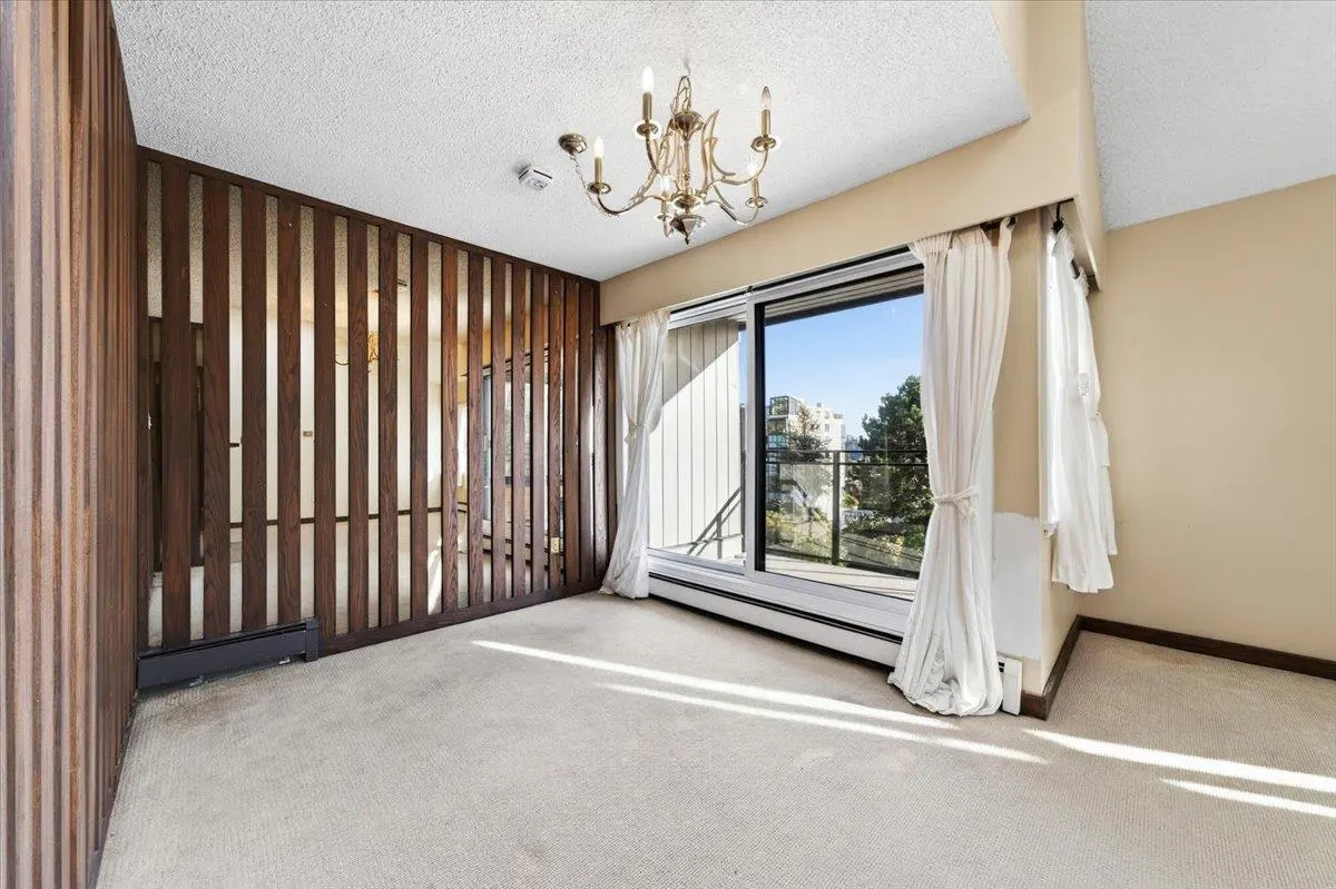 An empty room with beige carpet, walls, and a white ceiling. A brown wood slat wall, chandelier, and sliding glass door with white curtains are present.