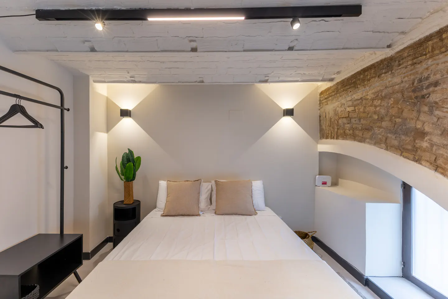 Bedroom with white walls, brick accent, and a bed with beige pillows. Black clothing rack and side table are visible.