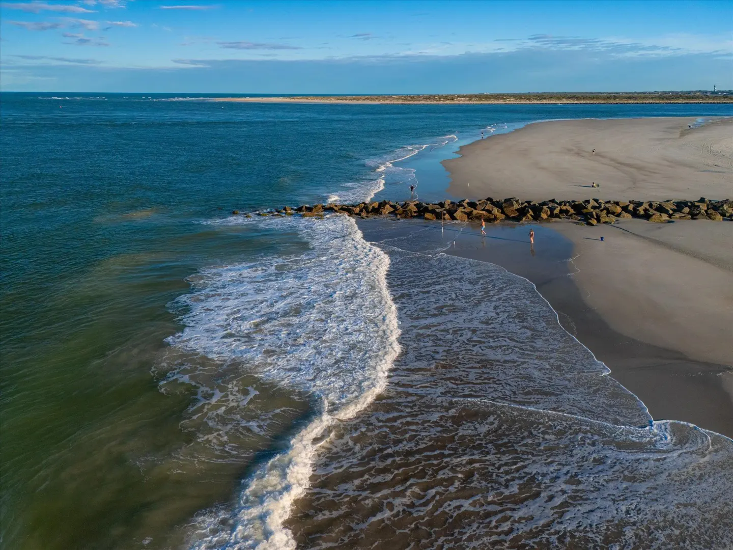 Aerial view of a sandy beach with a rock jetty extending into the blue ocean under a clear sky.