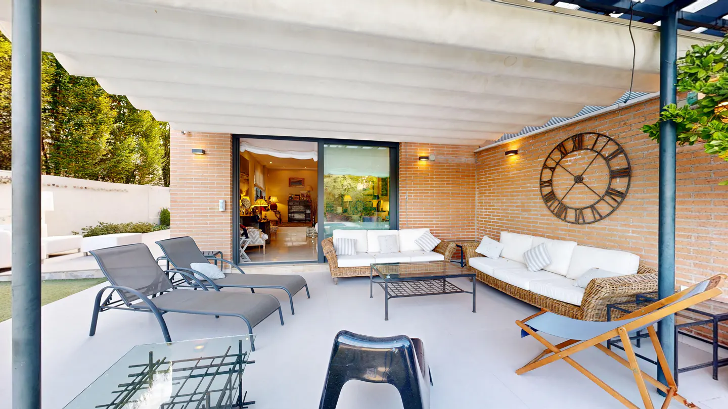 Outdoor patio with lounge chairs, wicker sofas, and a large clock on a brick wall. Sliding glass doors lead inside.