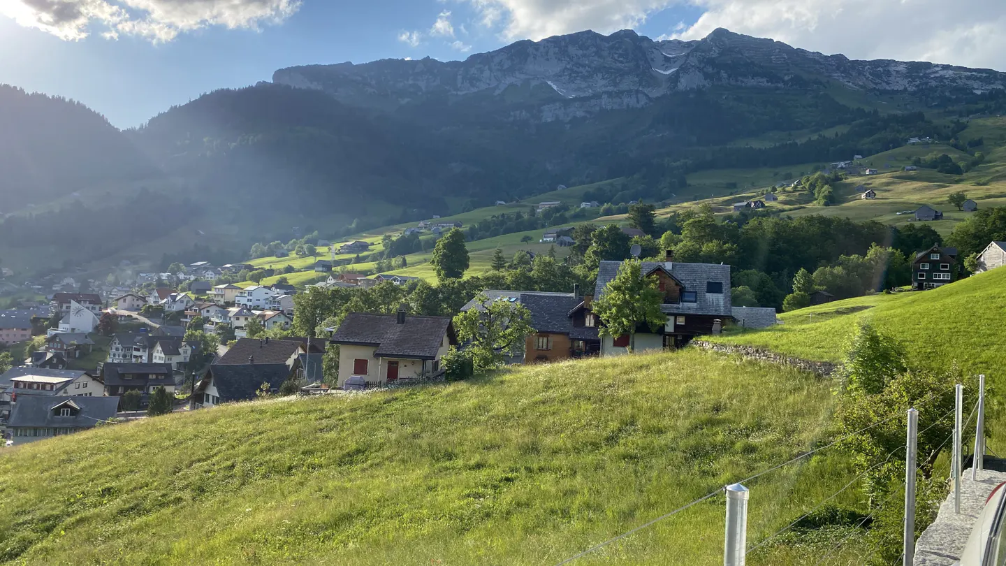 Scenic view of a village nestled in green hills, with mountains in the background under a partly cloudy sky.