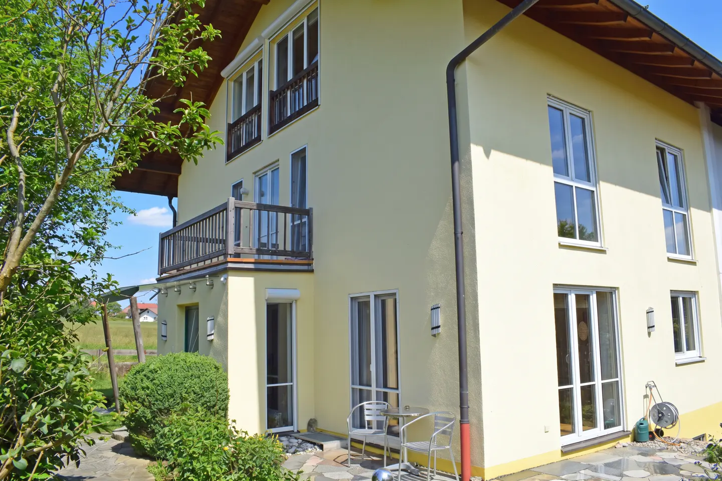 Two-story yellow house with a brown roof, white windows, and a wooden balcony. A small patio with chairs is visible.