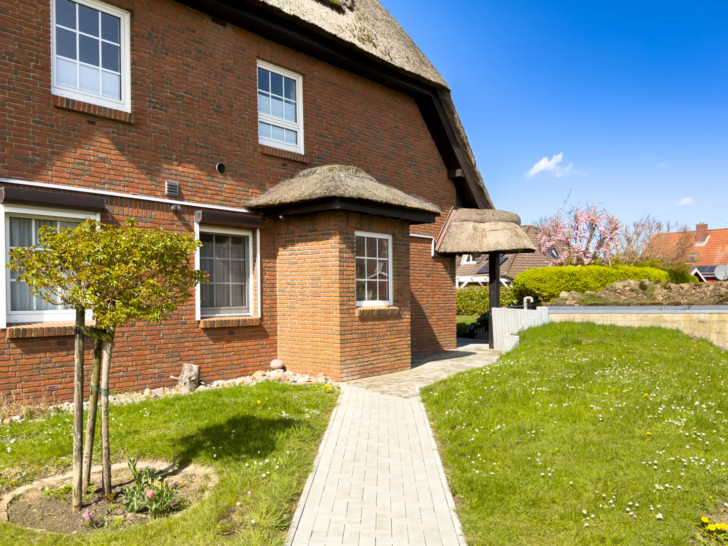 Brick house with a thatched roof and white windows. A stone path leads to the entrance, surrounded by green grass and a small tree.