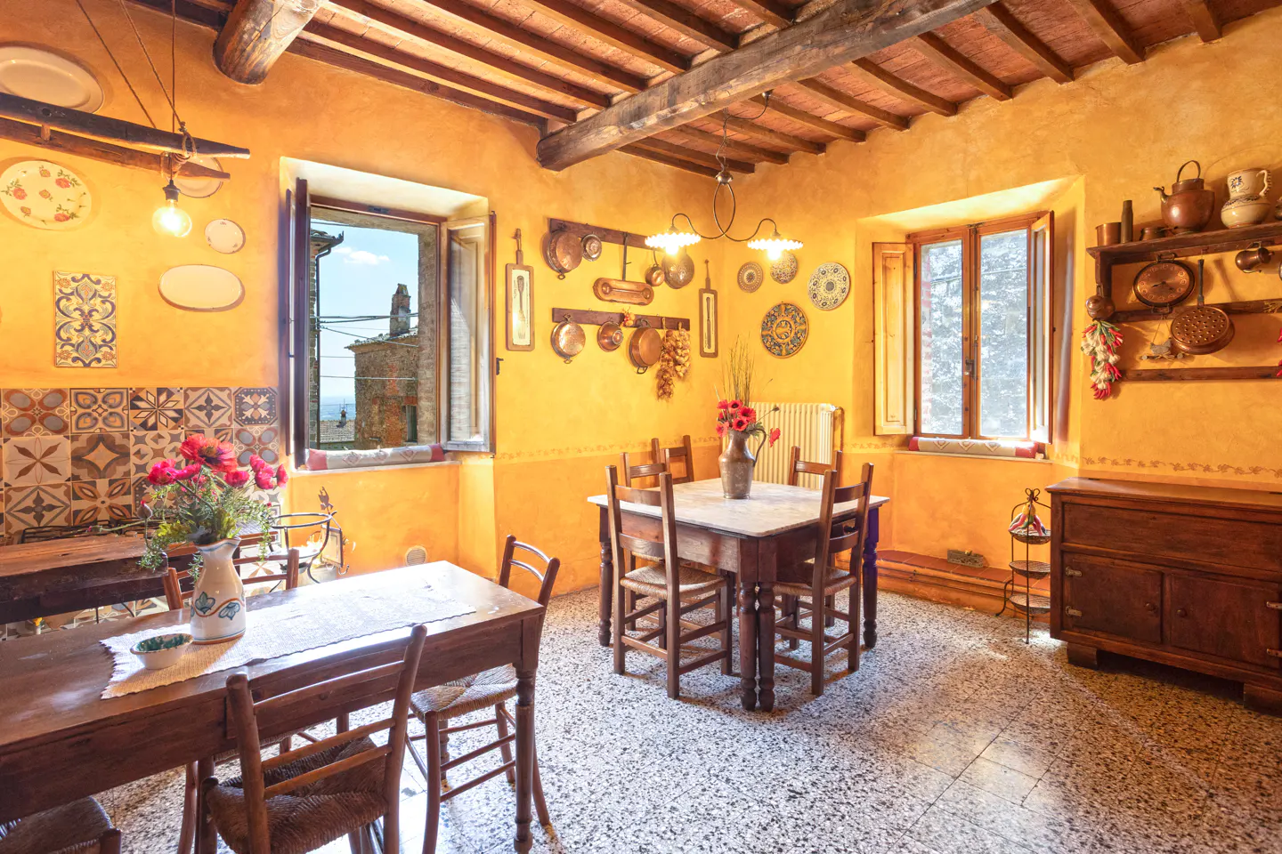 A rustic dining room with yellow walls, wooden tables and chairs, and decorative plates and copper pots on the walls. Windows offer a view of the outside.